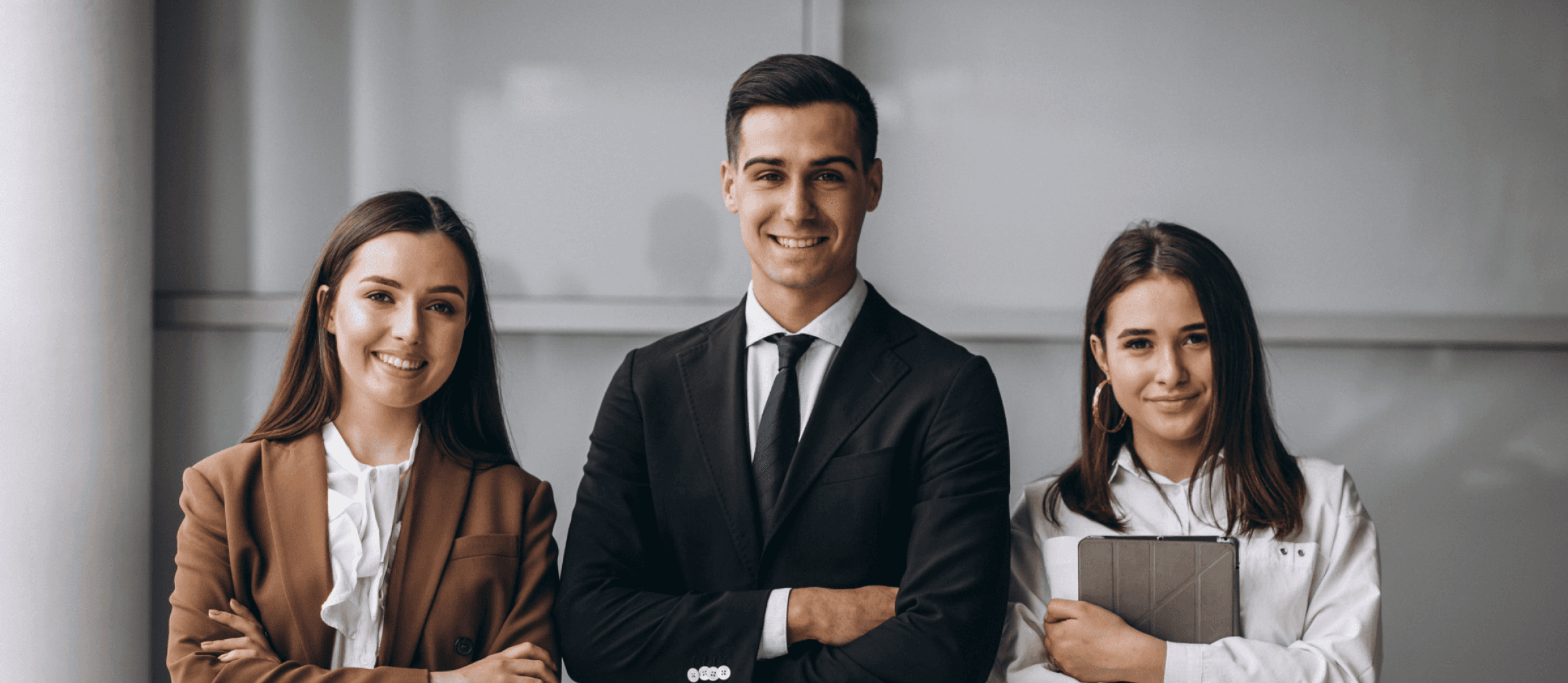 Three professionally dressed individuals stand side by side, confidently smiling towards the camera, embodying teamwork and diversity in a modern business setting.