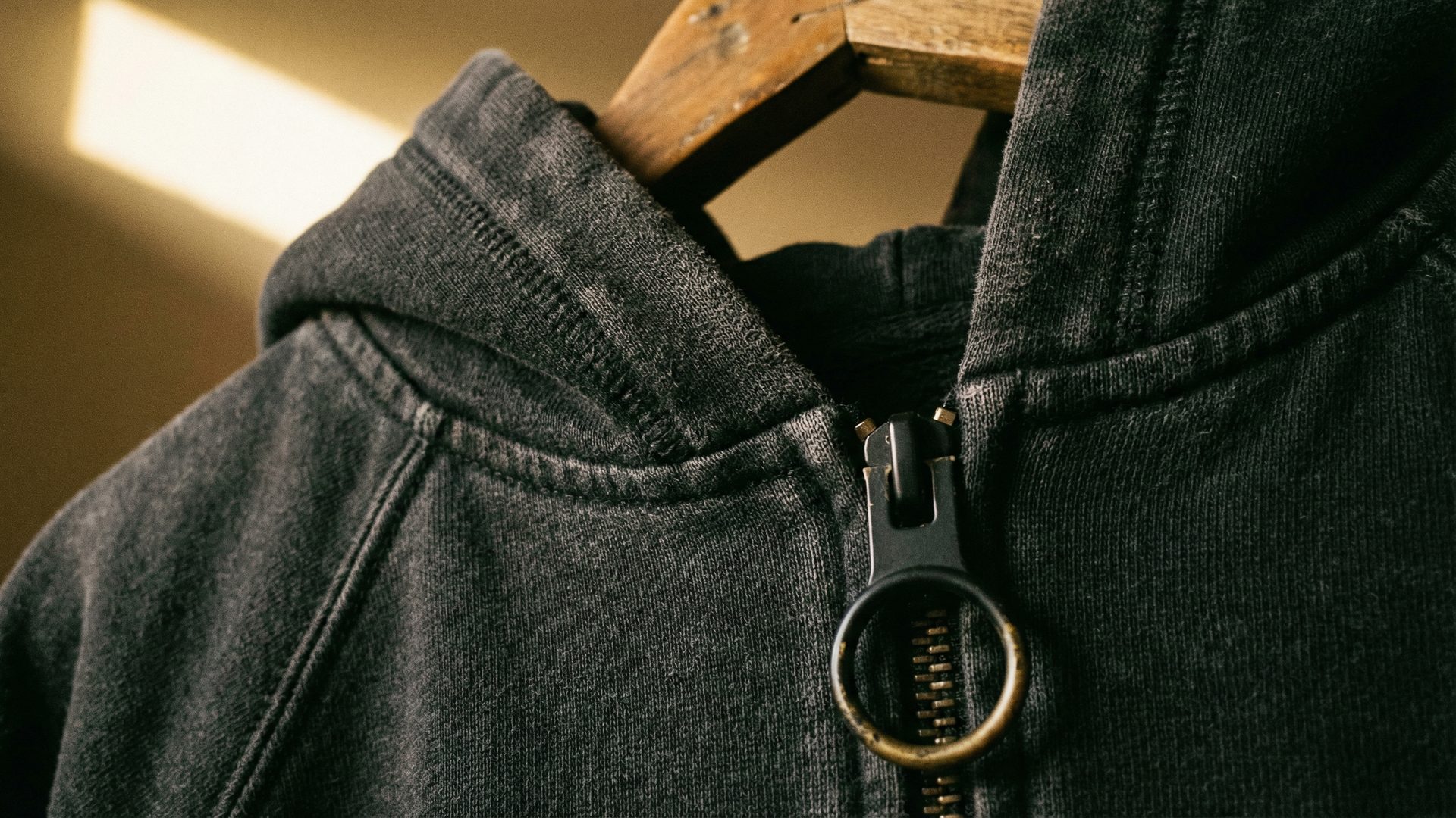 Extreme close-up of a premium heavyweight washed-black hoodie draped over a dark wooden hanger, showing the rich texture of the garment-dyed fleece, the quality of the flat-locked seams, and a custom matte black metal zipper pull.