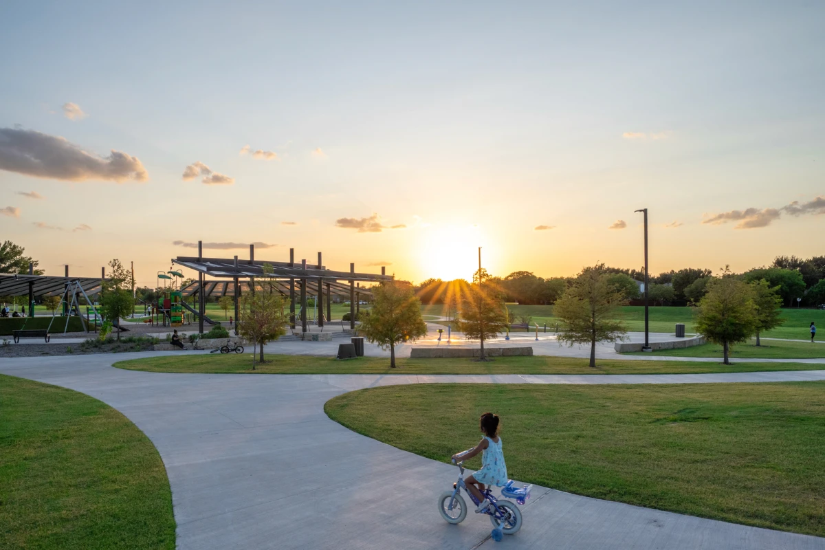 Child riding a bike at sunset through a community park with curved paths and playground shade canopy designed by East Fork