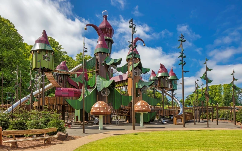 A large, imaginative outdoor adventure playground surrounded by trees and greenery. The structure features wooden towers, bridges, slides, and climbing elements, designed with a fairytale theme inspired by oversized leaves, mushrooms, and whimsical pointed rooftops painted in green, red, and brown. A wide open path and grassy area run alongside the play space, with benches placed nearby. The bright blue sky and lush surroundings create a playful, storybook atmosphere.