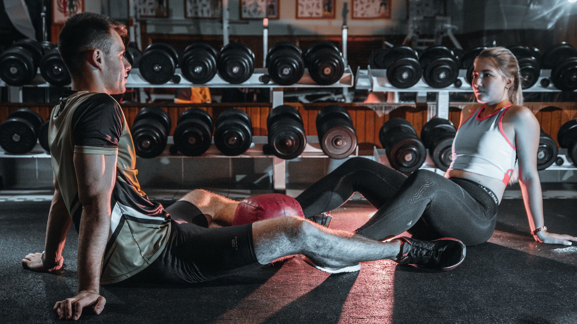 A man and woman sit on a gym floor facing each other and talking, with a dumbbell rack in the background.