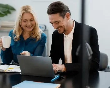 Businessman and woman collaborating on laptop, discussing work with notebook and coffee cup nearby together.