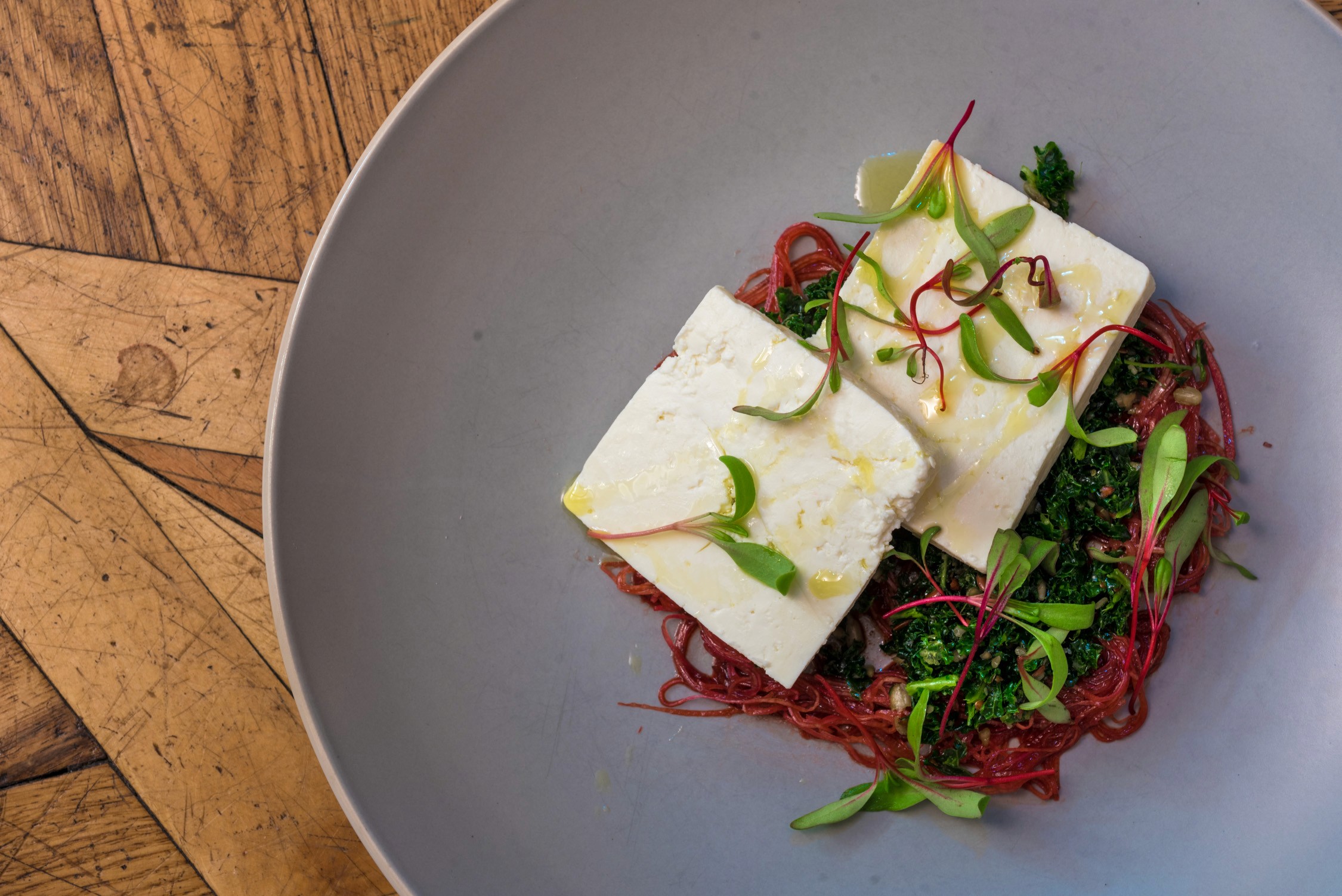 Two rectangular pieces of white tofu topped with green onions and red chili peppers over a bed of beet noodles in a gray