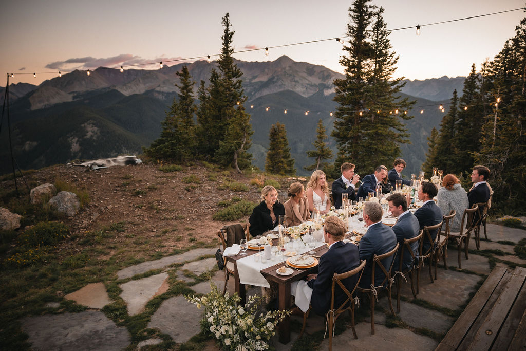 Wedding guests seated at a long outdoor dinner table in the mountains