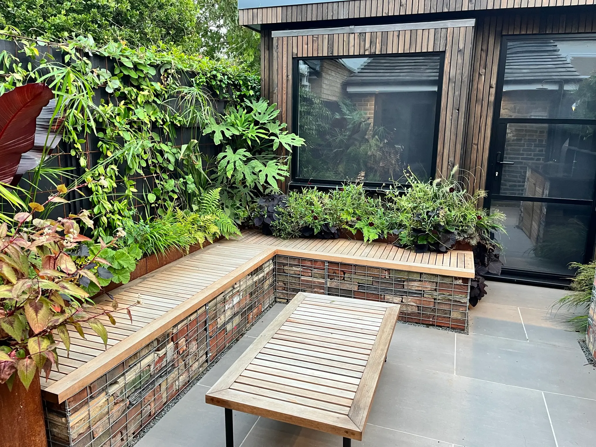 A cozy outdoor space featuring a stone bench, wooden table, and lush greenery surrounding a modern building.