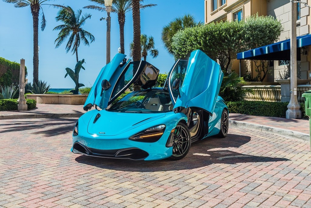 A stunning blue McLaren 720S with dihedral doors open, parked in a luxurious setting with palm trees and an elegant building in the background. This exotic supercar features a sleek design, powerful twin-turbo V8 engine, and unparalleled performance, making it the perfect choice for a luxury car rental experience.
