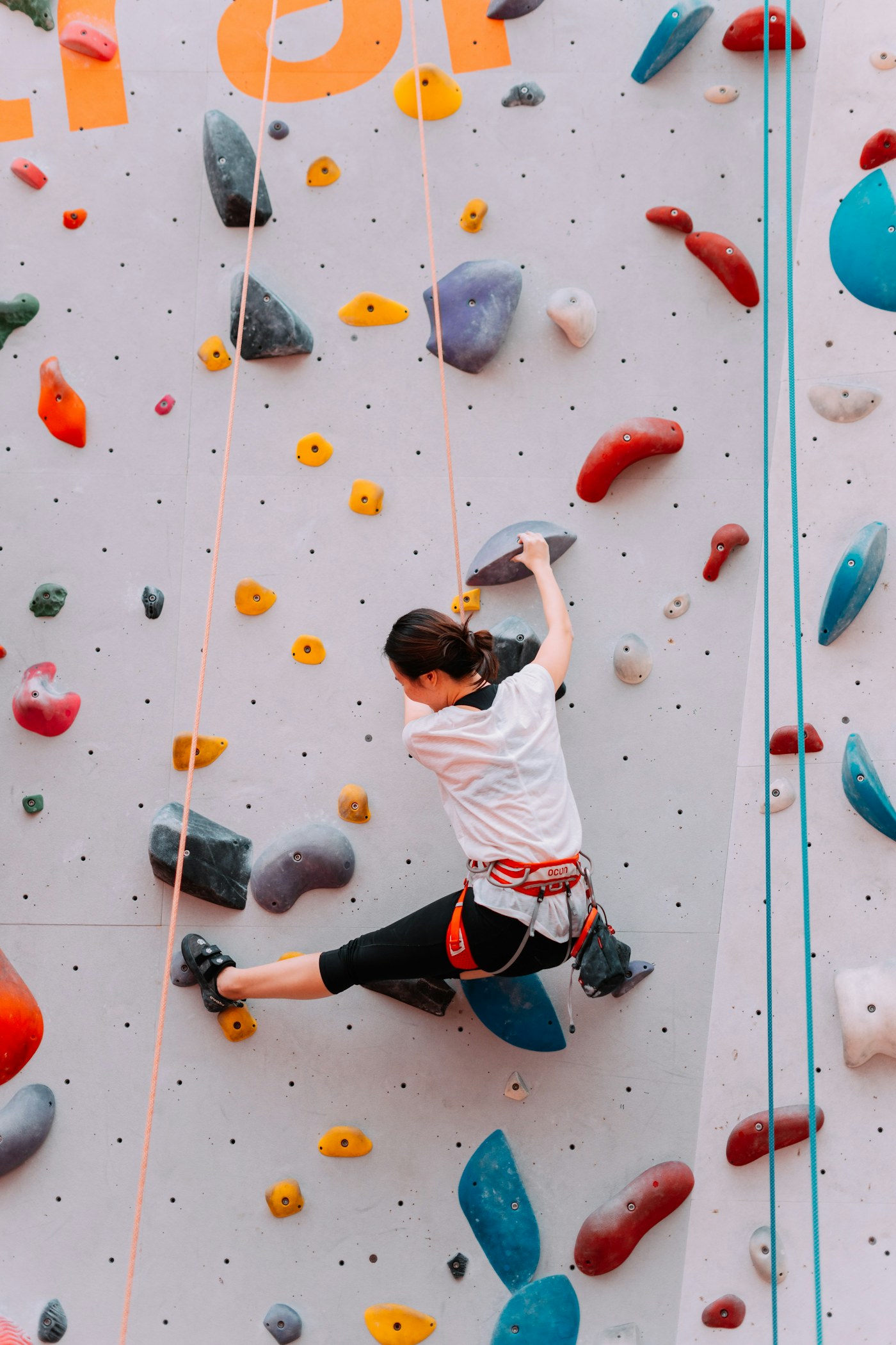 A kid in a white T-shirt climbs a pale wall covered in red, blue and yellow holds