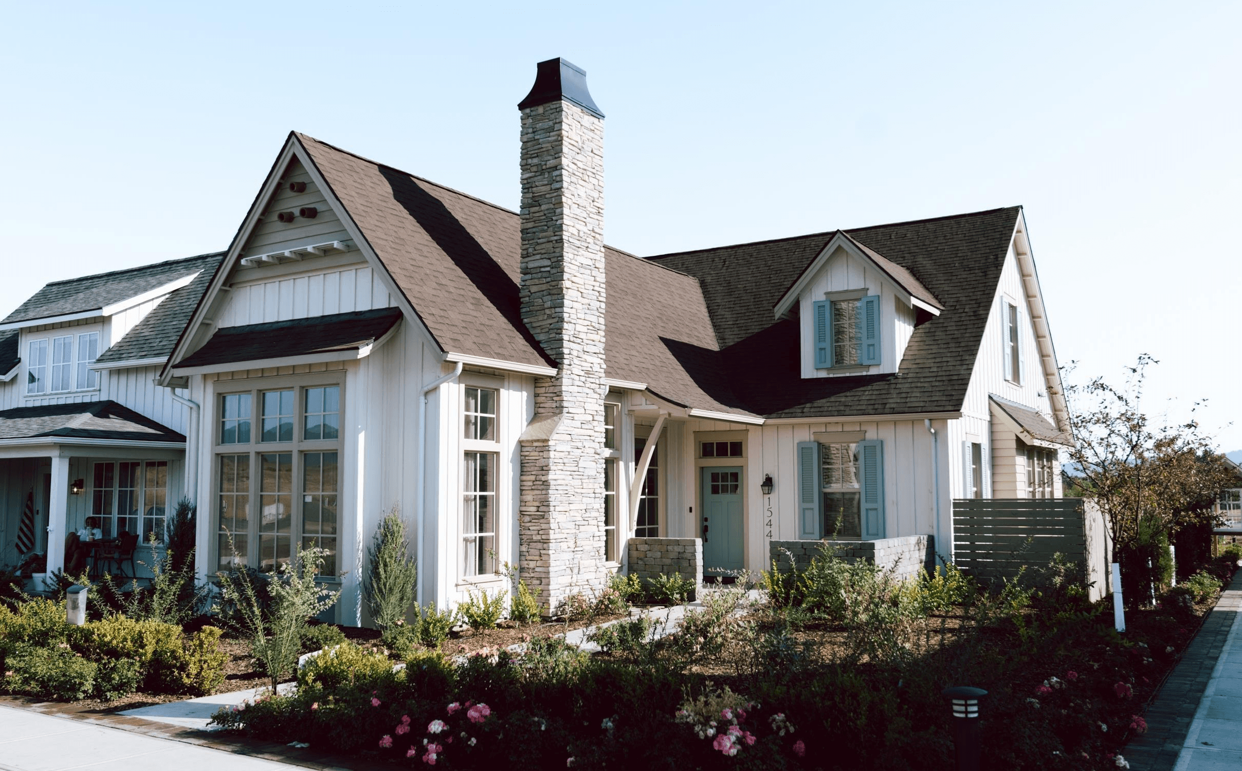 Cream cottage with teal shutters, a brown roof, a stone chimney, and landscaping at Woodbury.