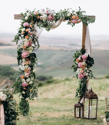 Floral archway adorned with greenery and blooms, set against a scenic landscape. Lanterns on either side.