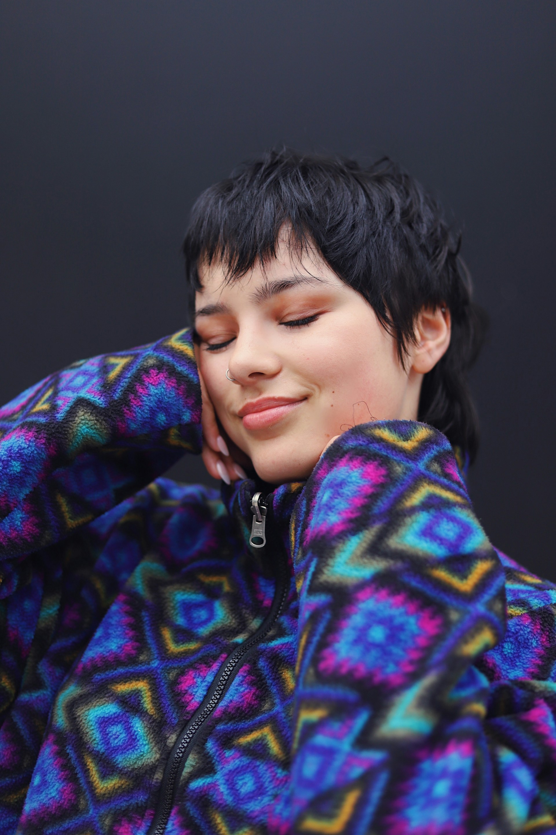 A close-up of a person with short, dark shaggy hair and eyes closed, smiling gently while resting their chin on their hands. They are wearing a vibrant, geometric-patterned fleece jacket.