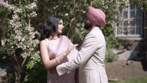 Man in an off-white suit with a pink turban and woman in a pink dress holding each other and gazing at each other in front of a tree and a historic building.