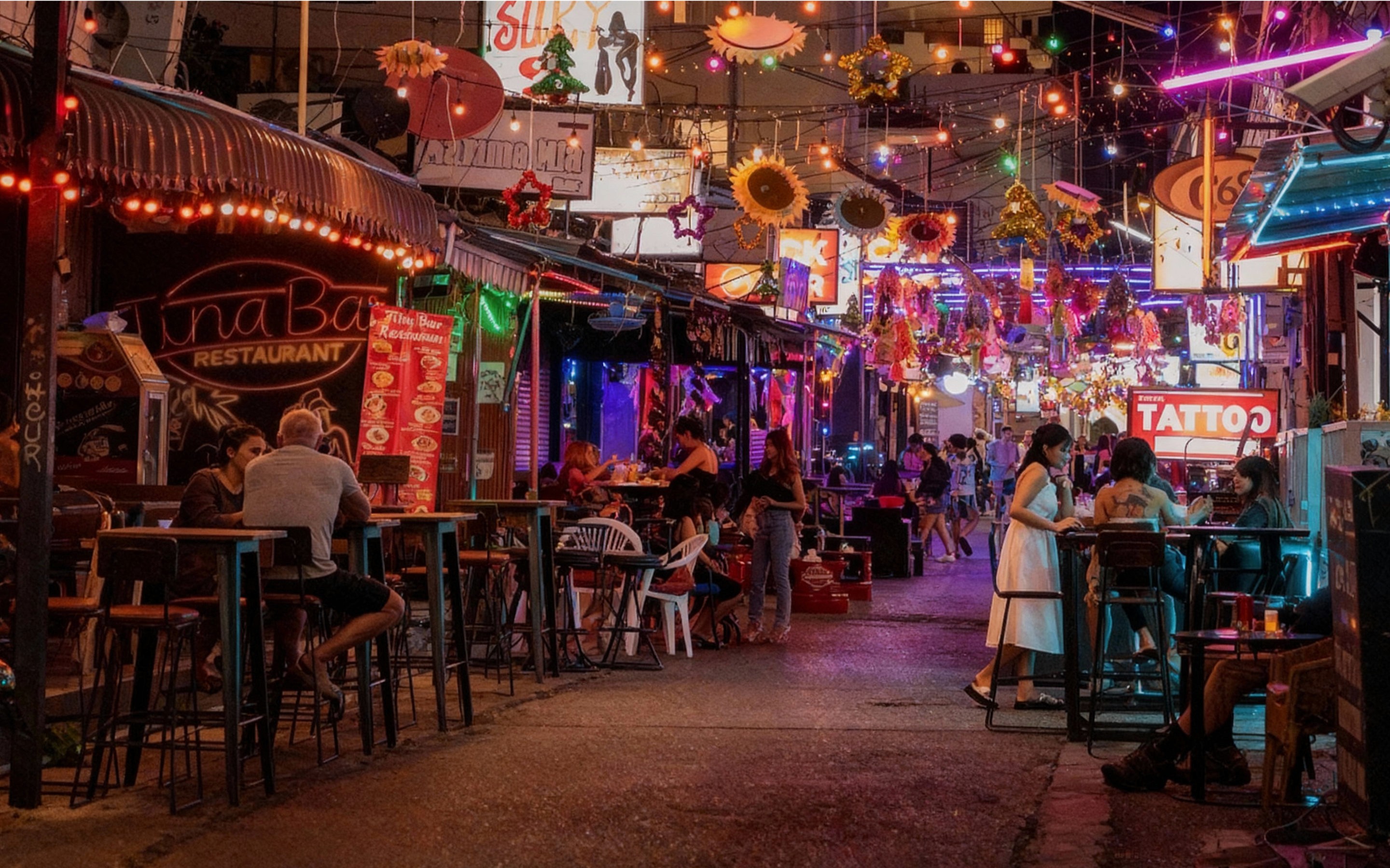Lively Soi Bintabaht in Hua Hin at dusk, featuring a bustling street with neon-lit bars, pool tables, and people socializing, framed by palm trees and colorful signs under a twilight sky.