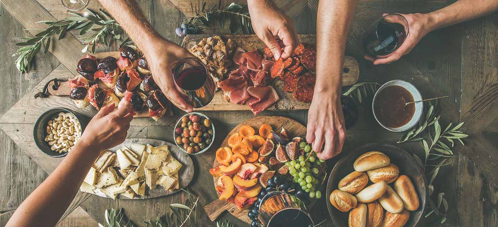 Hands reaching for charcuterie board with cheese, fruit, meats, bread, olives, and wine.
