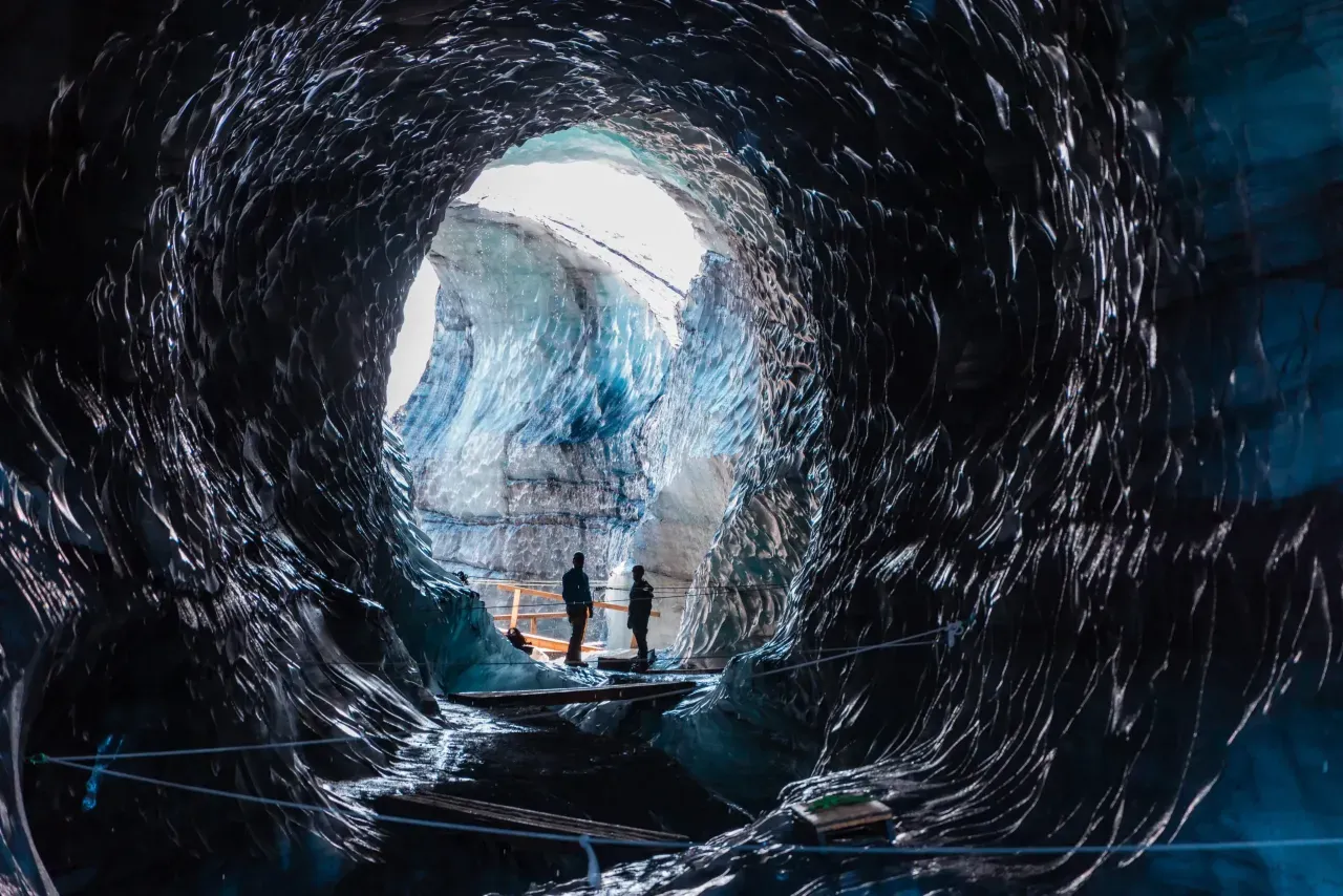 Looking out from inside the dark, textured ice tunnels of Katla Ice Cave, with silhouettes of people standing at the entrance