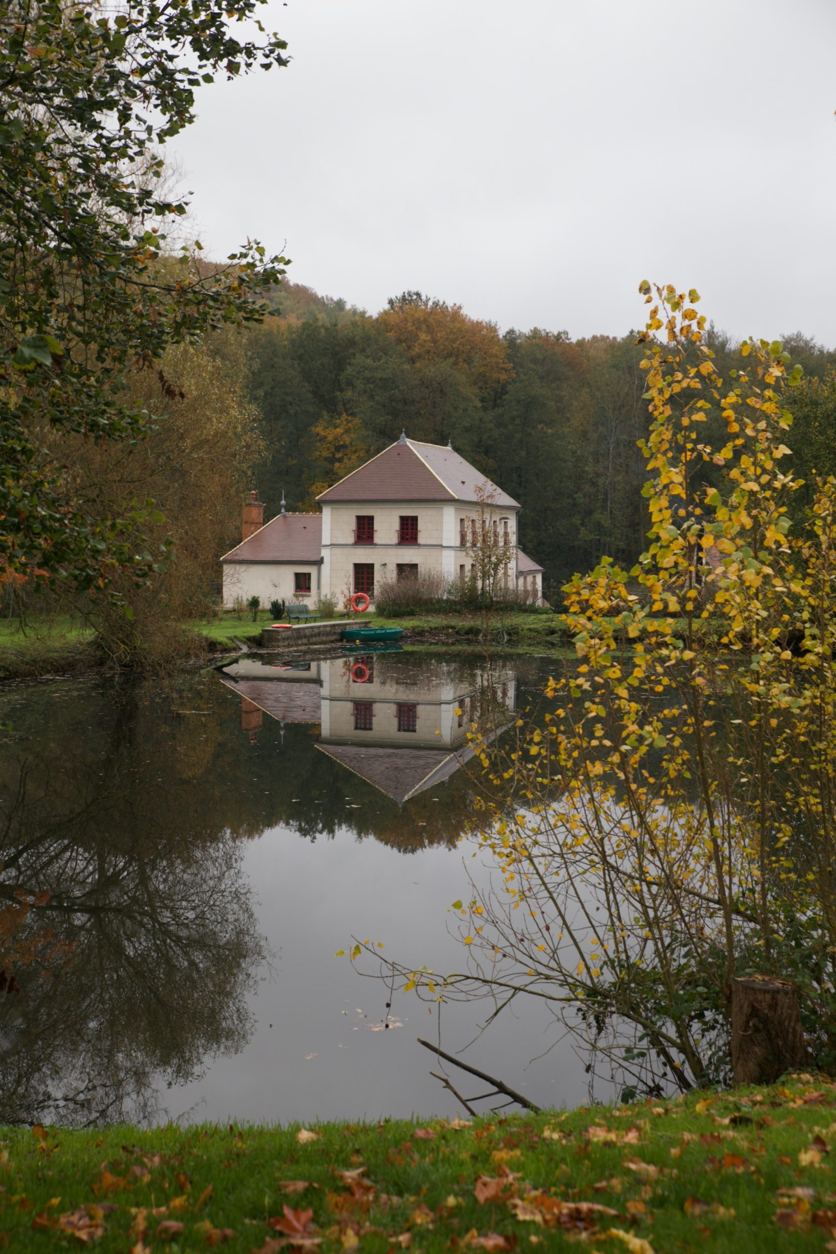 White country manor house with a mansard roof reflected in a still pond, surrounded by autumn trees