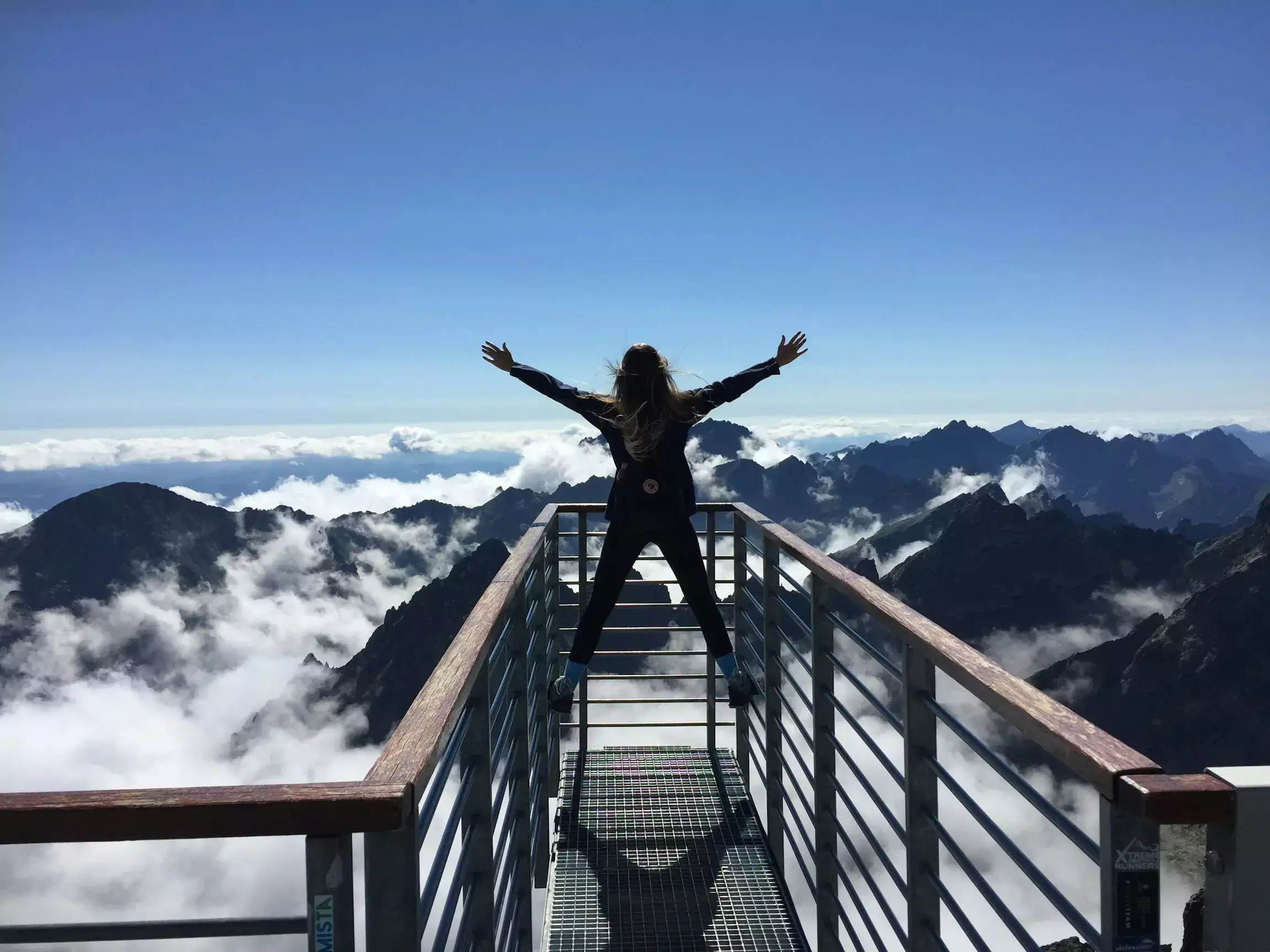 a woman standing at an observation point in a celebratory posture with hands above her head while staring out into the mountains and a blue sky