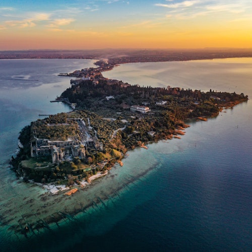 Aerial view of a narrow peninsula with lush greenery and buildings, surrounded by blue water at sunset.