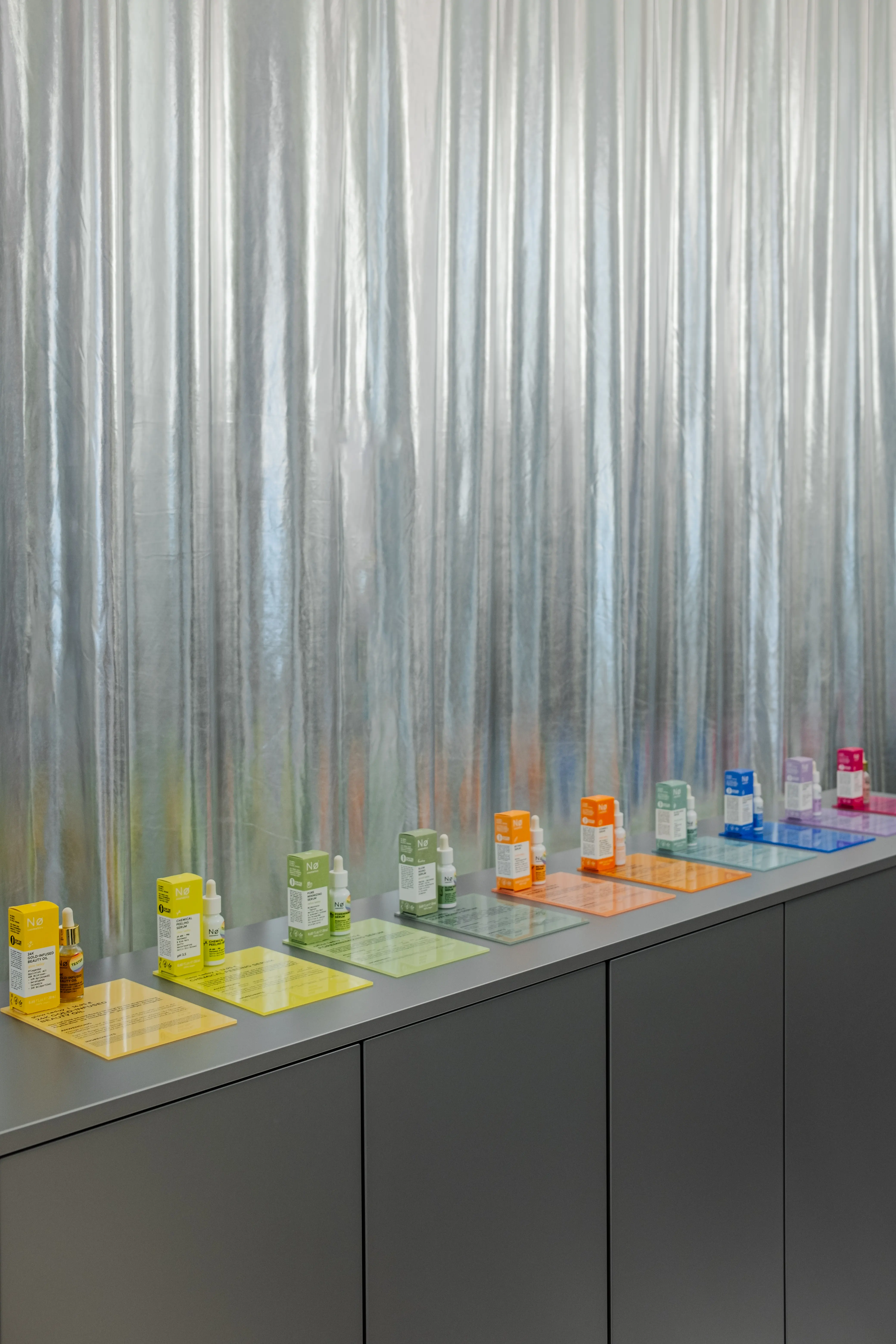 Skincare products on colorful acrylic mats in a row on the grey sideboard in front of a shimmering curtain.