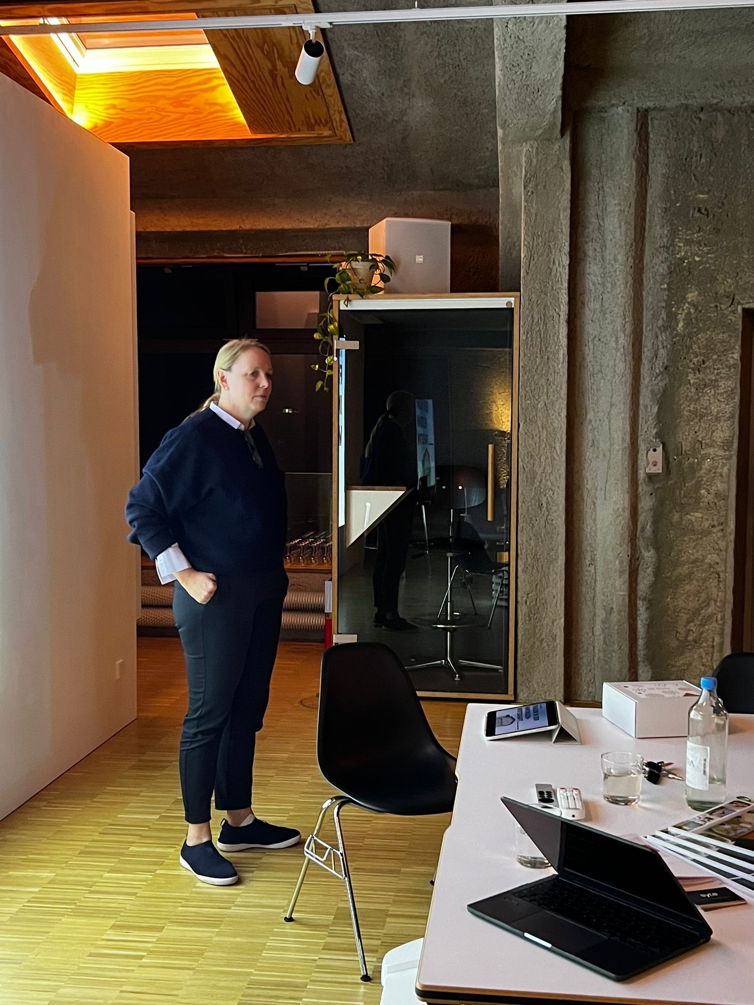 Person steht in einem modernen Innenraum mit Sichtbeton und Holzfußboden; im Hintergrund eine kleine Glaskabine und ein Schreibtisch mit technischen Geräten.