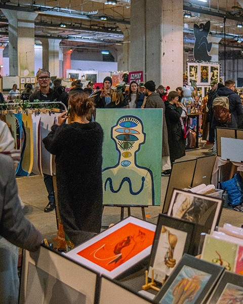 Zdjęcie przedstawiające artystkę na pierwszym planie na stanowisku targowym. W tle duża grupa ludzi przeglądająca stanowiska.