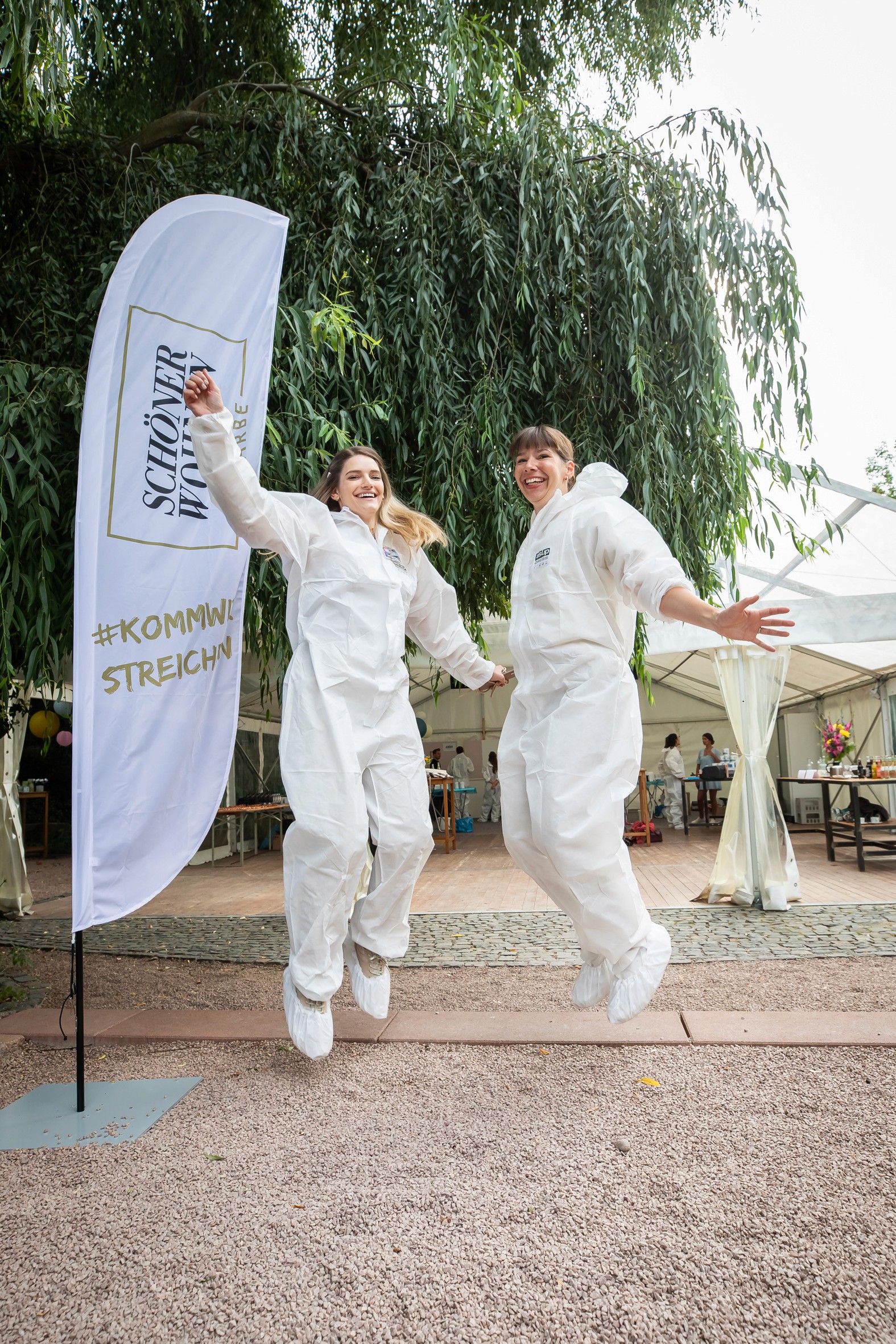 Zwei Teilnehmer:innen springen in Schutzanzügen bei einem Kreativ-Workshop von Schöner Wohnen Farbe im Freien.
