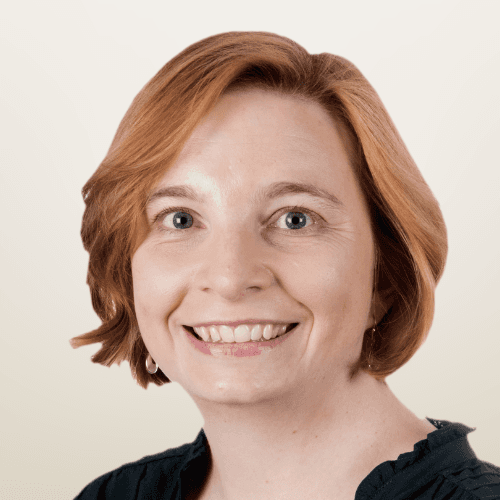 A man with glasses and a light gray blazer smiles against a softly lit background.