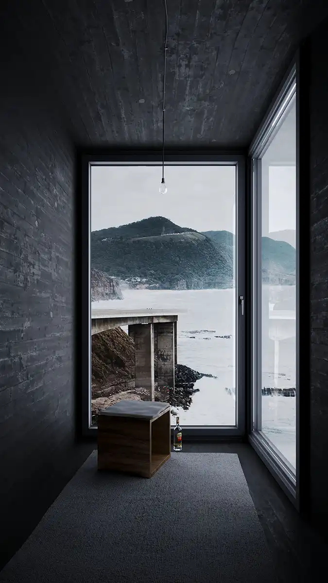 Minimalist concrete room with large panoramic window overlooking dramatic coastal landscape, wooden stool, and bottle. Contemplative architectural interior.