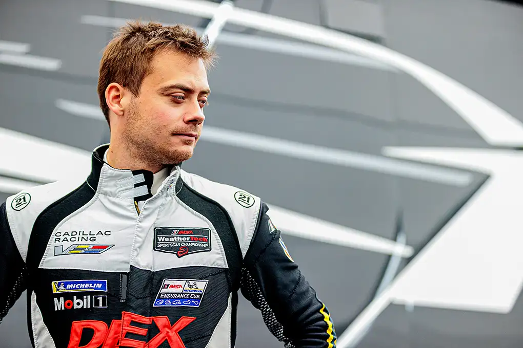 Cadillac Racing driver, Louis Delétraz, stands confidently in front of a gray patterned wall, looking to the right.