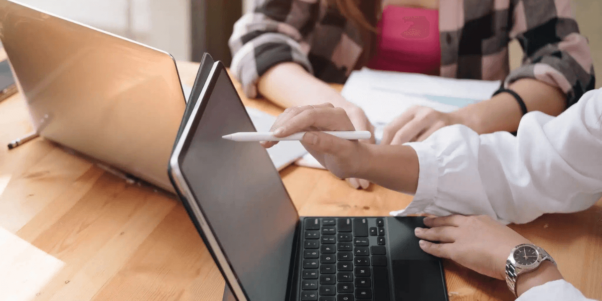 Two people at a wooden table use laptops, one pointing with a stylus at a tablet. The scene conveys collaboration and focus in a work setting.