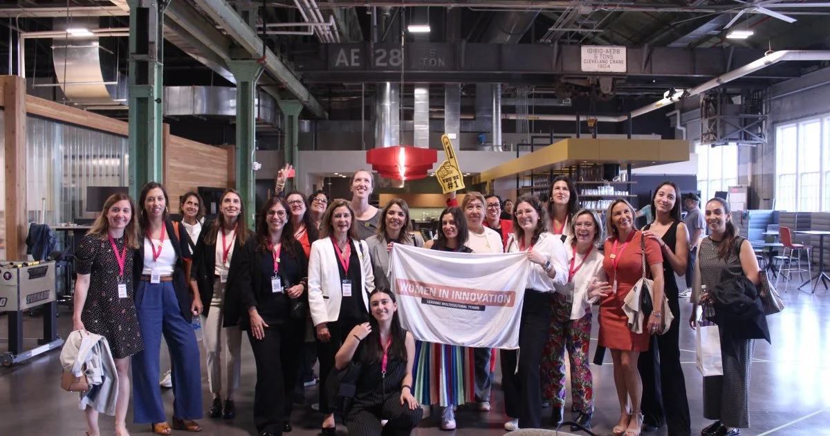 A diverse group of people gathers in a spacious industrial setting, some holding signs, smiling for a group photo.