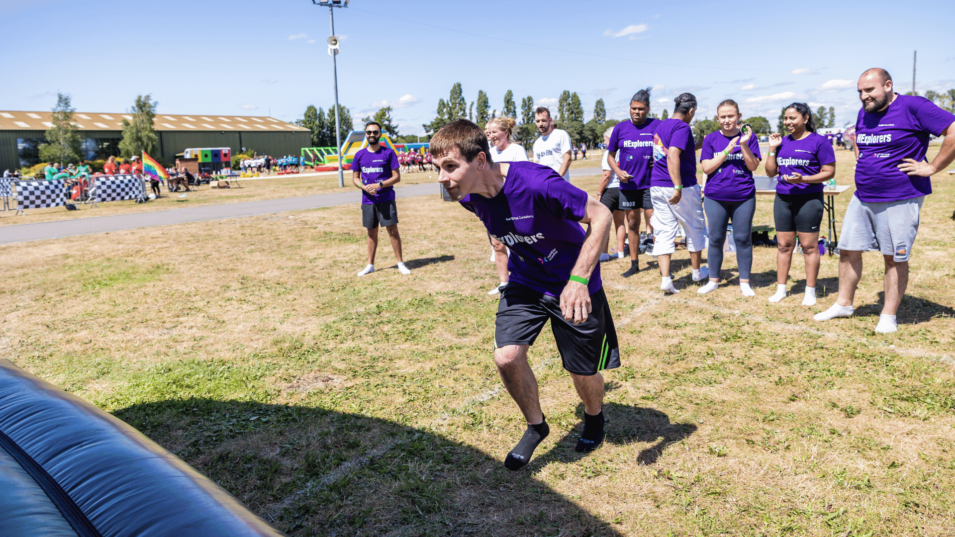 Outdoor team-building event with participants wearing matching blue shirts, taking part in inflatable obstacle courses and games, smiling and cheering, with the words 'ENTER A TEAM' in large bold white and green text across the image.