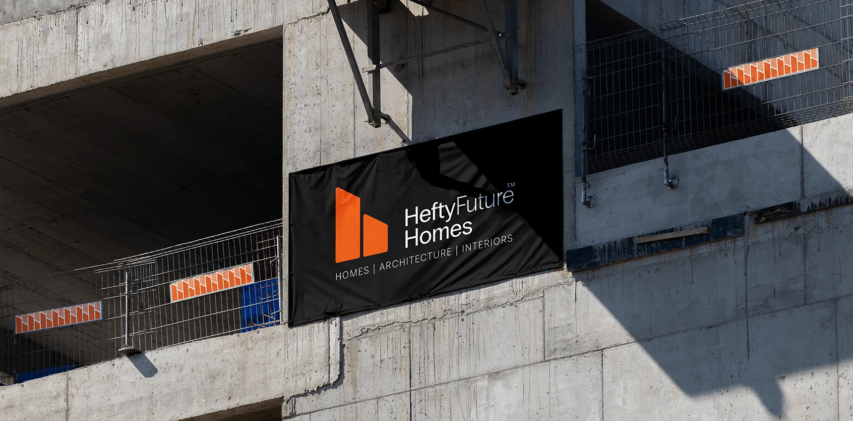 A black banner on a bare concrete building under construction, showing the orange monogram, "HeftyFuture Homes" and "Homes | Architecture | Interiors", with orange pattern panels on the safety railings.