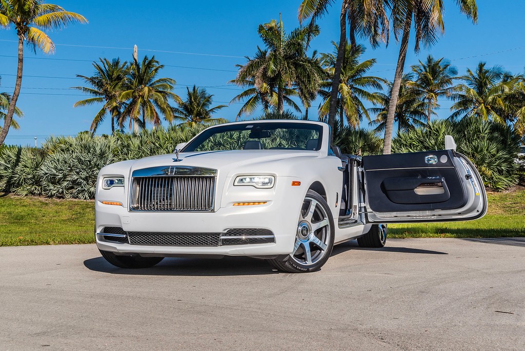  Front view of a white Rolls-Royce Dawn with open doors, parked in a tropical setting with palm trees in the background.