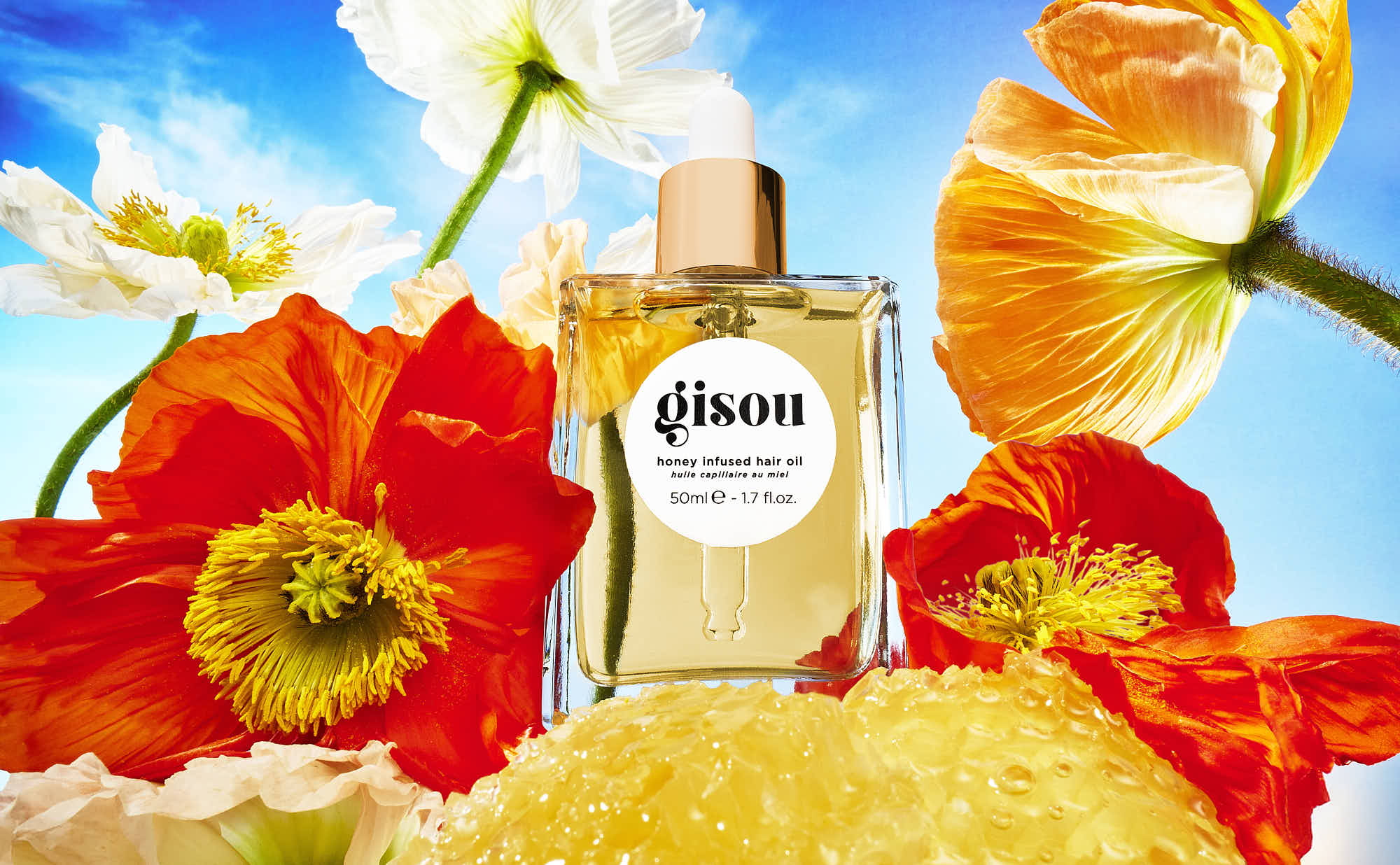 A bottle of oiled surrounded by colorful flowers against a bright blue sky.