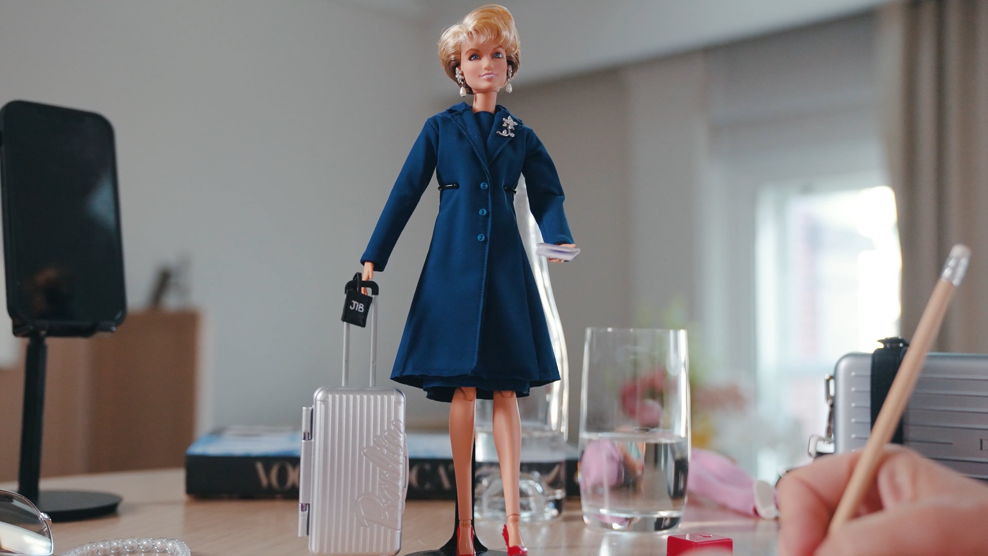 Barbie doll in a navy coat beside a small suitcase, arranged on a desk