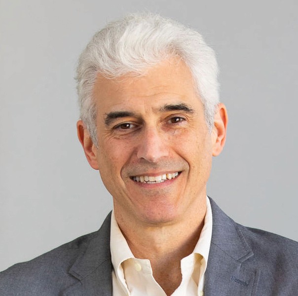 A smiling man with white hair, wearing a gray jacket and light shirt, against a neutral background.