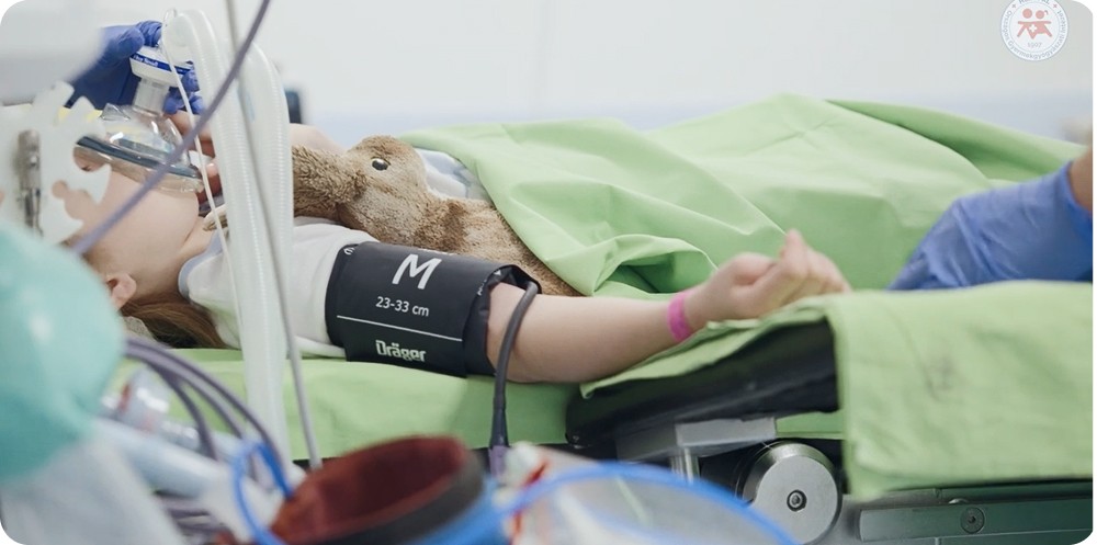 Altatásban fekvő kisgyermek kórházi ágyon, oxigénmaszkkal és plüssjátékkal.