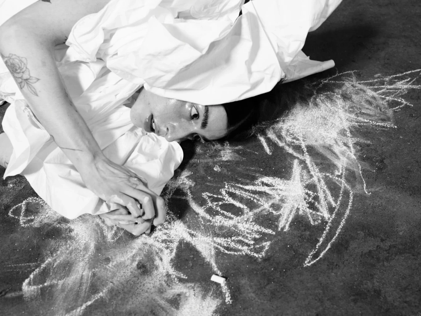 A woman in a white shirt lying on a dark floor surrounded by chalk drawings, her hands clasped beneath her cheek, a rose tattoo visible on her arm, black and white