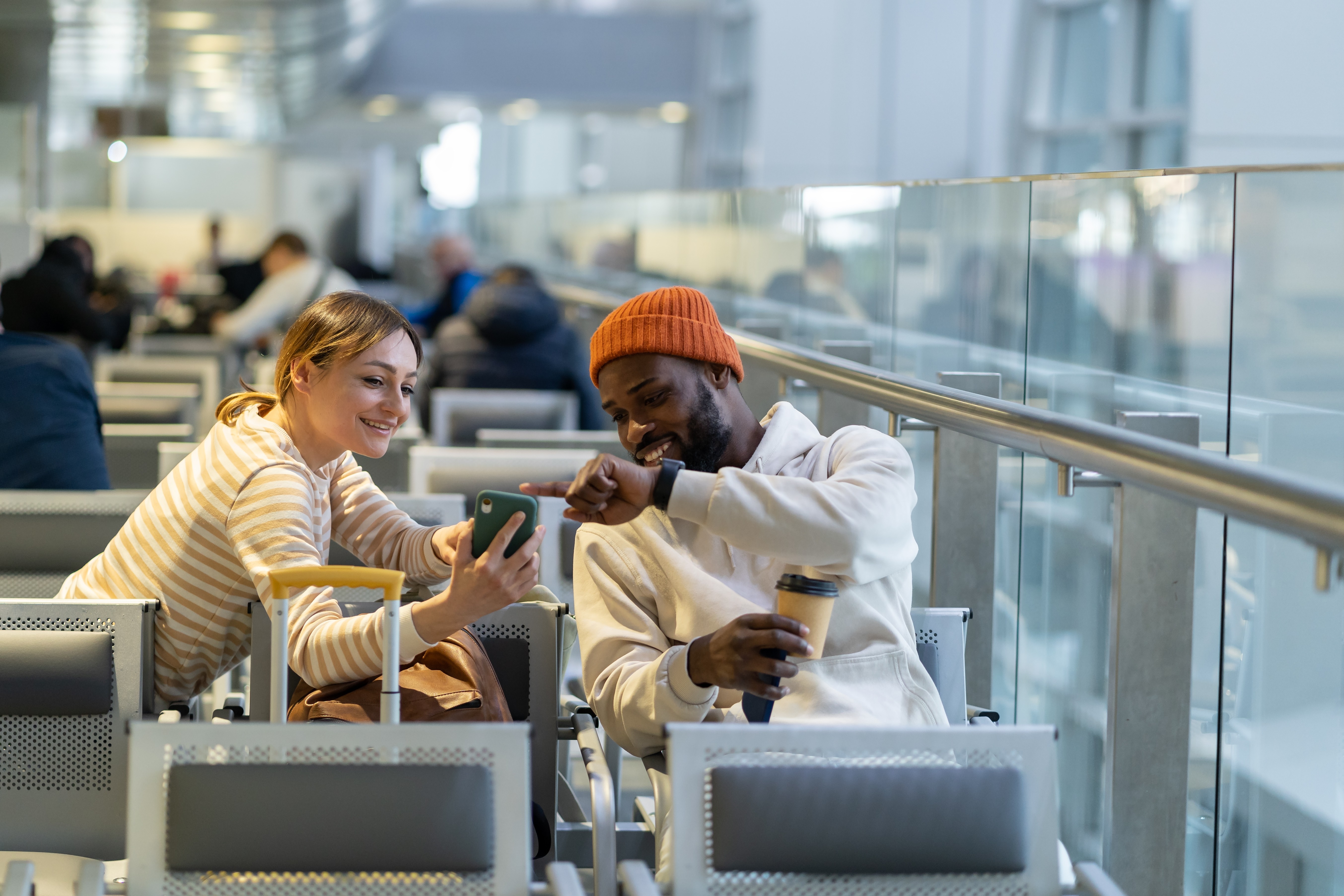 A photo of a couple sat in an airport, chatting while pointing at a mobile phone