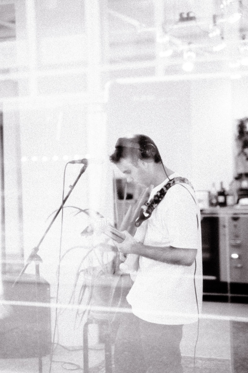 Black and white photo taken by Julia Roman during a live performance by Loveboy and His Imaginary Friends at the Collektiv in St.Gallen, Switzerland. The image captures a musician wearing headphones and playing an electric guitar, seen through a reflective glass. The atmospheric composition and soft focus evoke an intimate, dreamy concert setting.