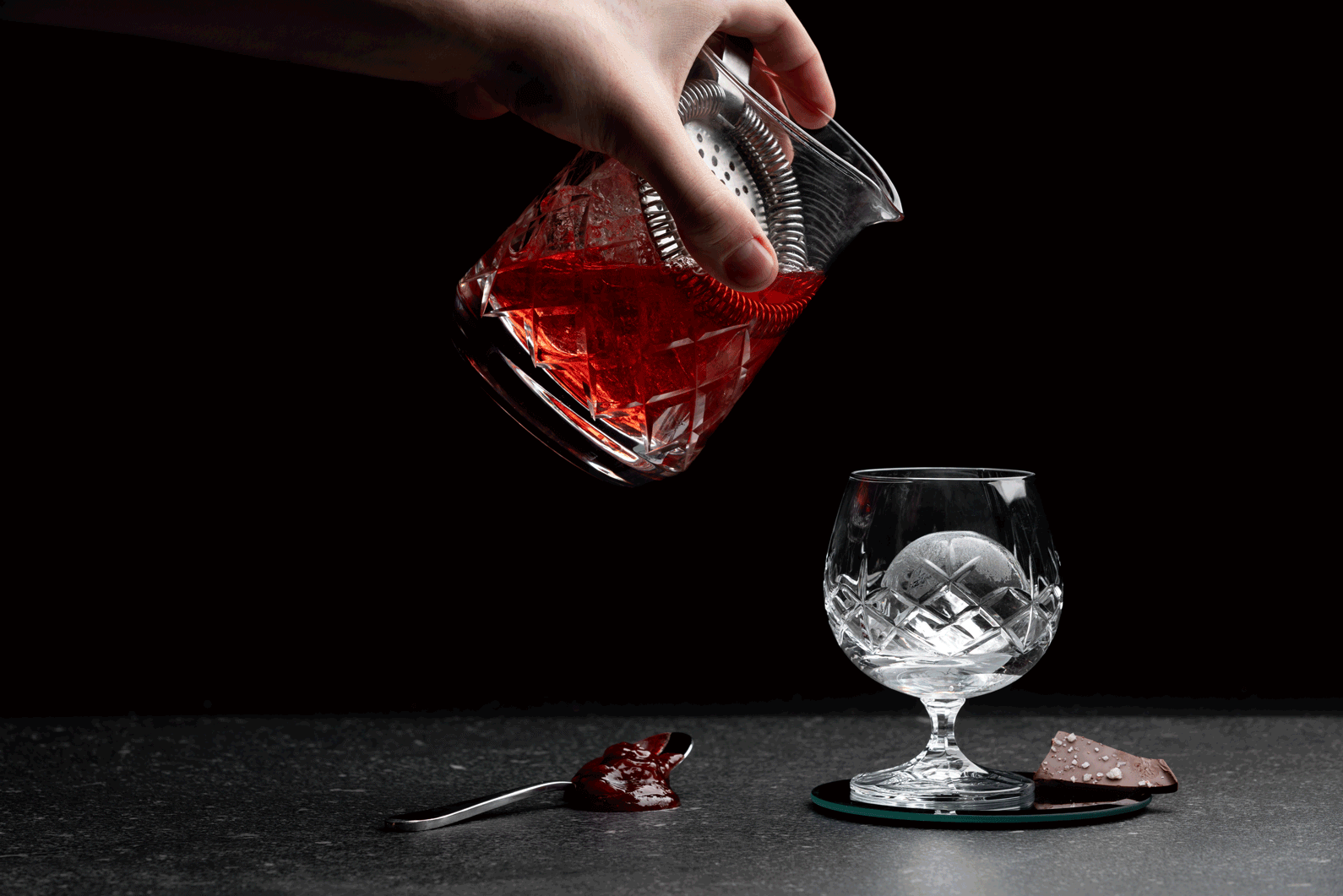 A hand elegantly pouring a deep red cocktail from a textured crystal mixing glass into a matching snifter glass with an ice sphere, set against a dark, moody background with gourmet chocolate and a spoon nearby.