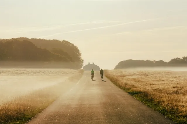 Two cyclists riding together through a misty rural field at sunrise