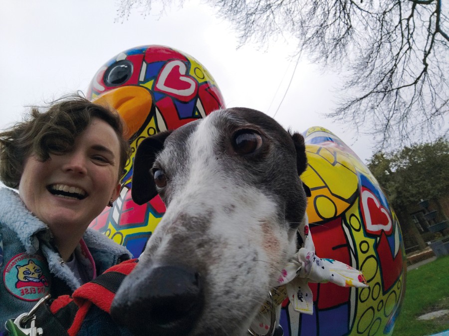 A photo of Amy, she is a white woman, with brown short hair. Her hair is a little curly and messy. She is laughing stood in front of large painted statue of a duck. She is next to her dog Eve, who is a greyhound. Eve's nose is super close to the camera and she has big dark eyes. She is black and white.