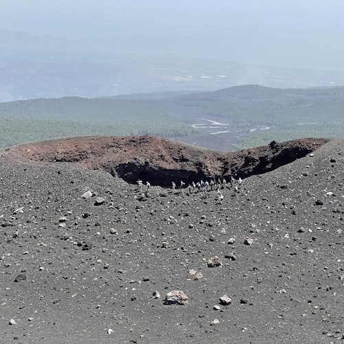 Um grupo de pessoas está em pé perto da borda de uma grande cratera vulcânica, com vistas distantes de colinas e do mar.