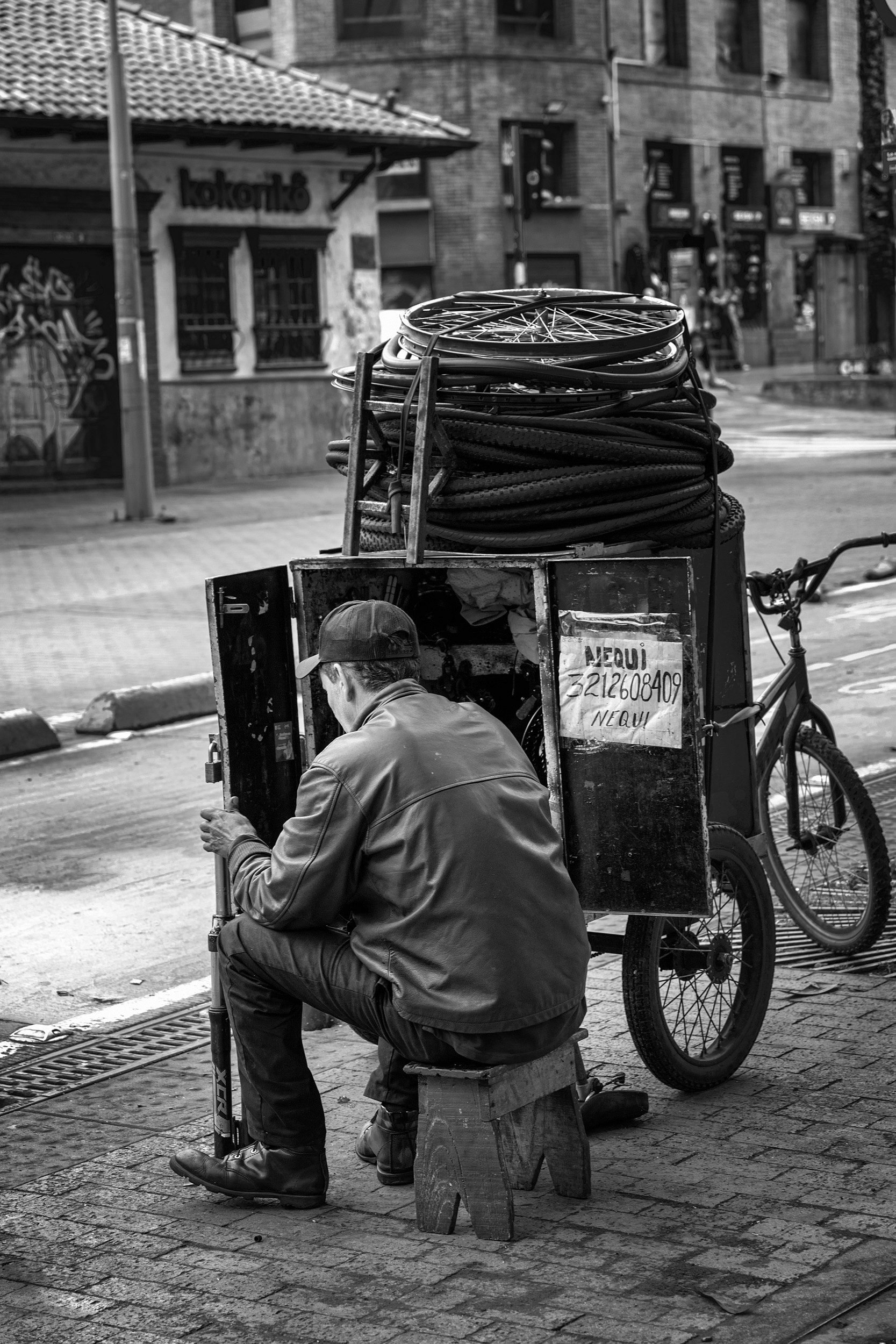 A bicycle mechanic sits on a small wooden stool, working quietly behind his mobile repair cart stacked with tires and parts, on a Bogotá sidewalk.