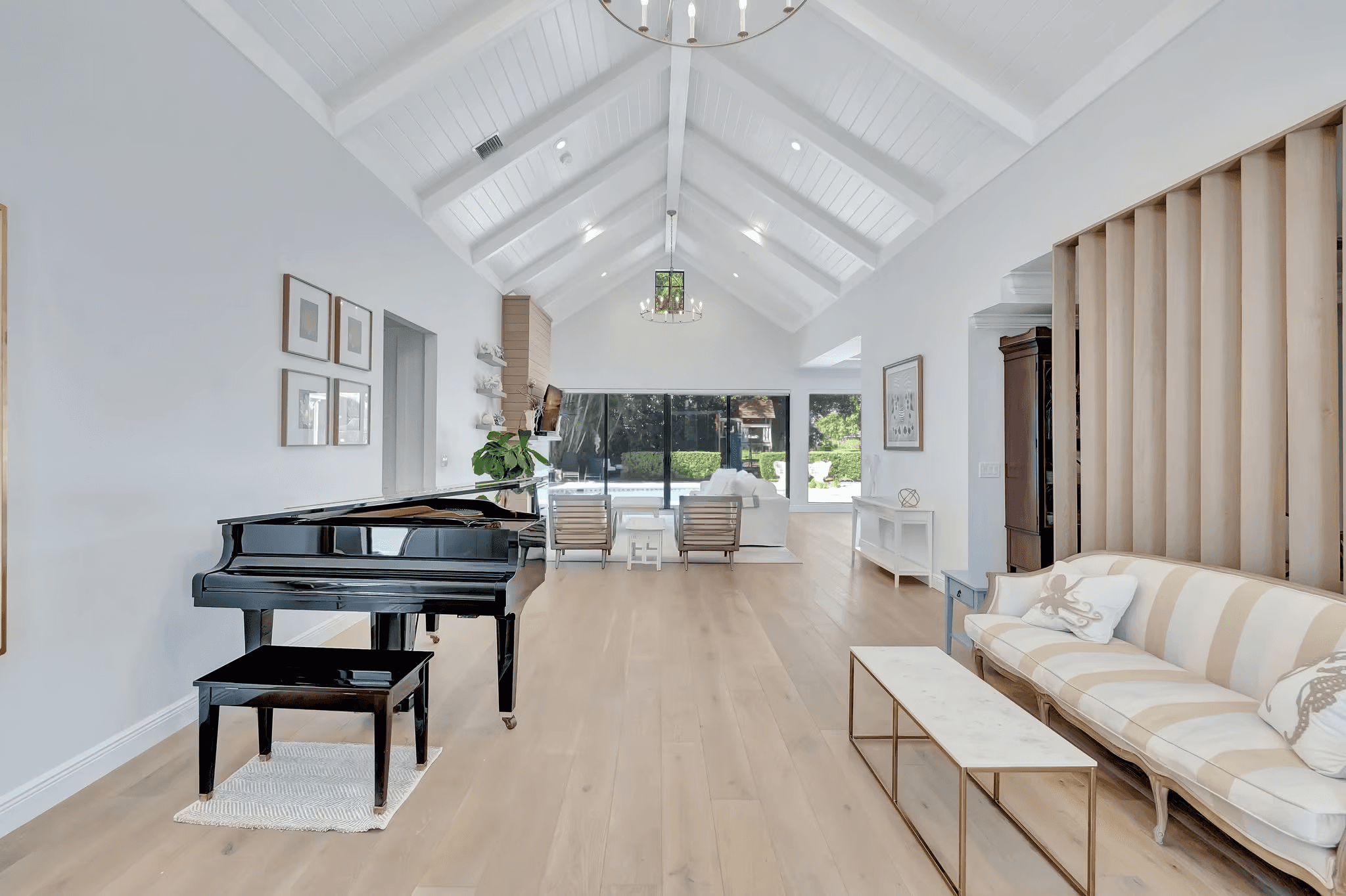 Elegant living room with cathedral ceiling, white beadboard, light oak floors, grand piano, and sliding glass doors overlooking pool area