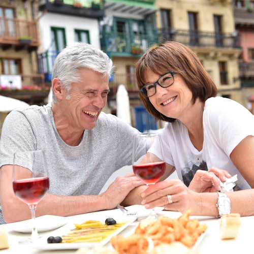 A smiling older couple with glasses enjoying a meal and drinks outdoors, with colorful buildings in the background.
