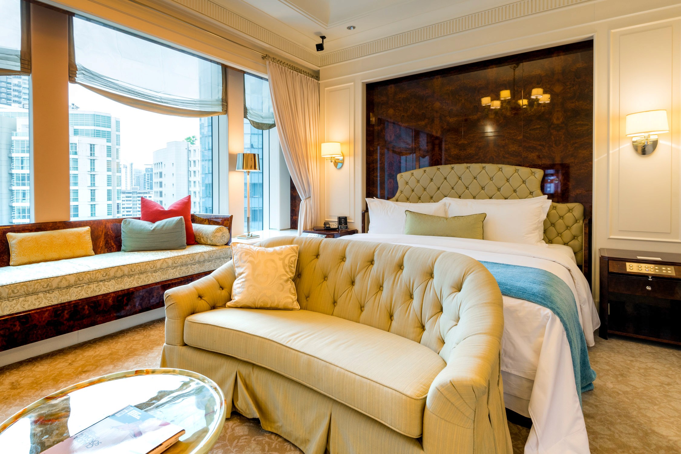 A luxury hotel room features a tufted cream sofa, a large bed with white linens, and a window overlooking a city.