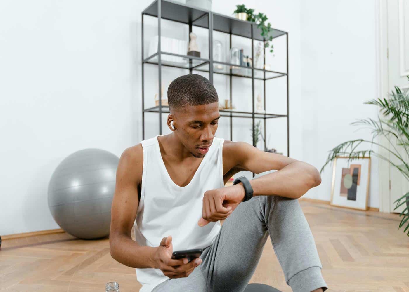 Man in workout clothes sitting on floor with phone in hand and looking at smartwatch