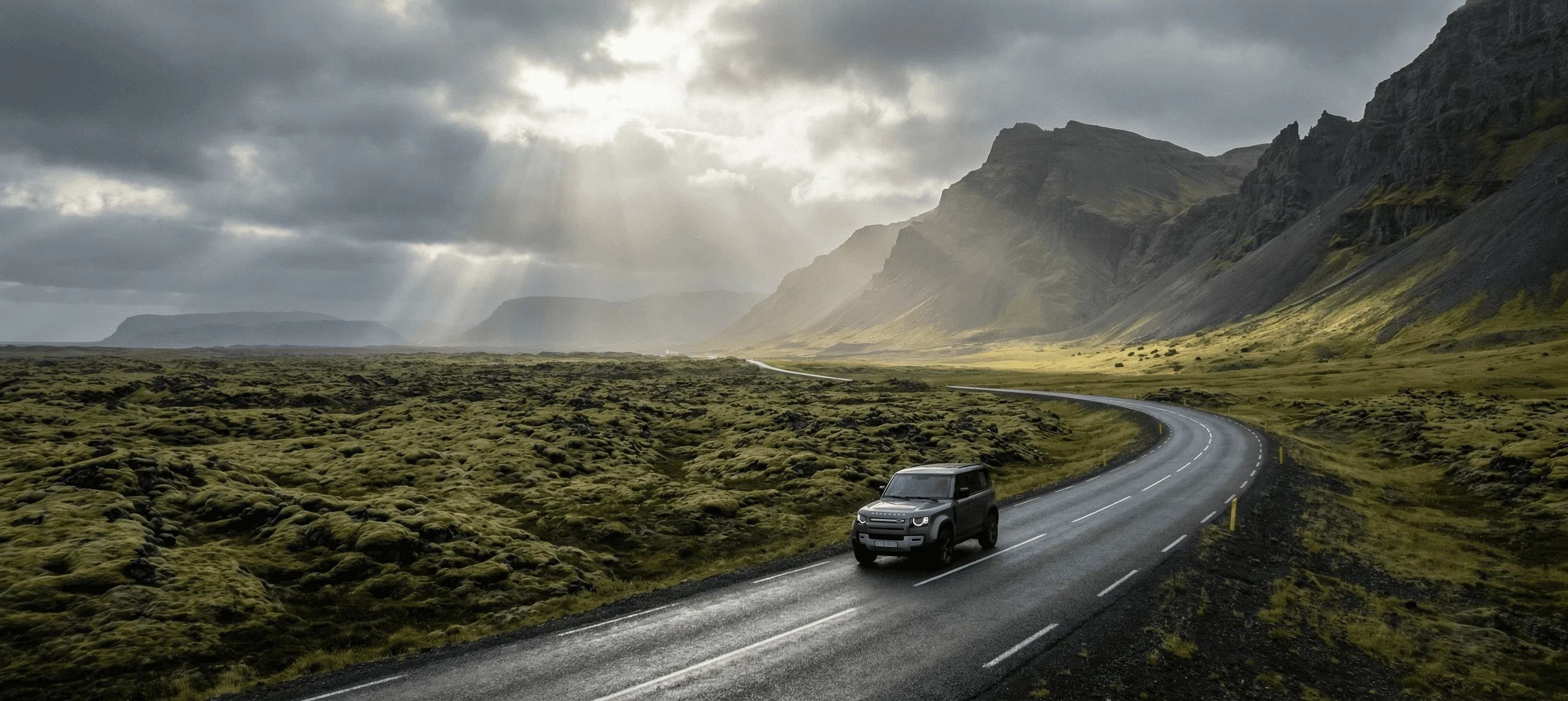 A grey SUV driving along a winding asphalt road through a green mossy valley with sun rays breaking through the clouds.